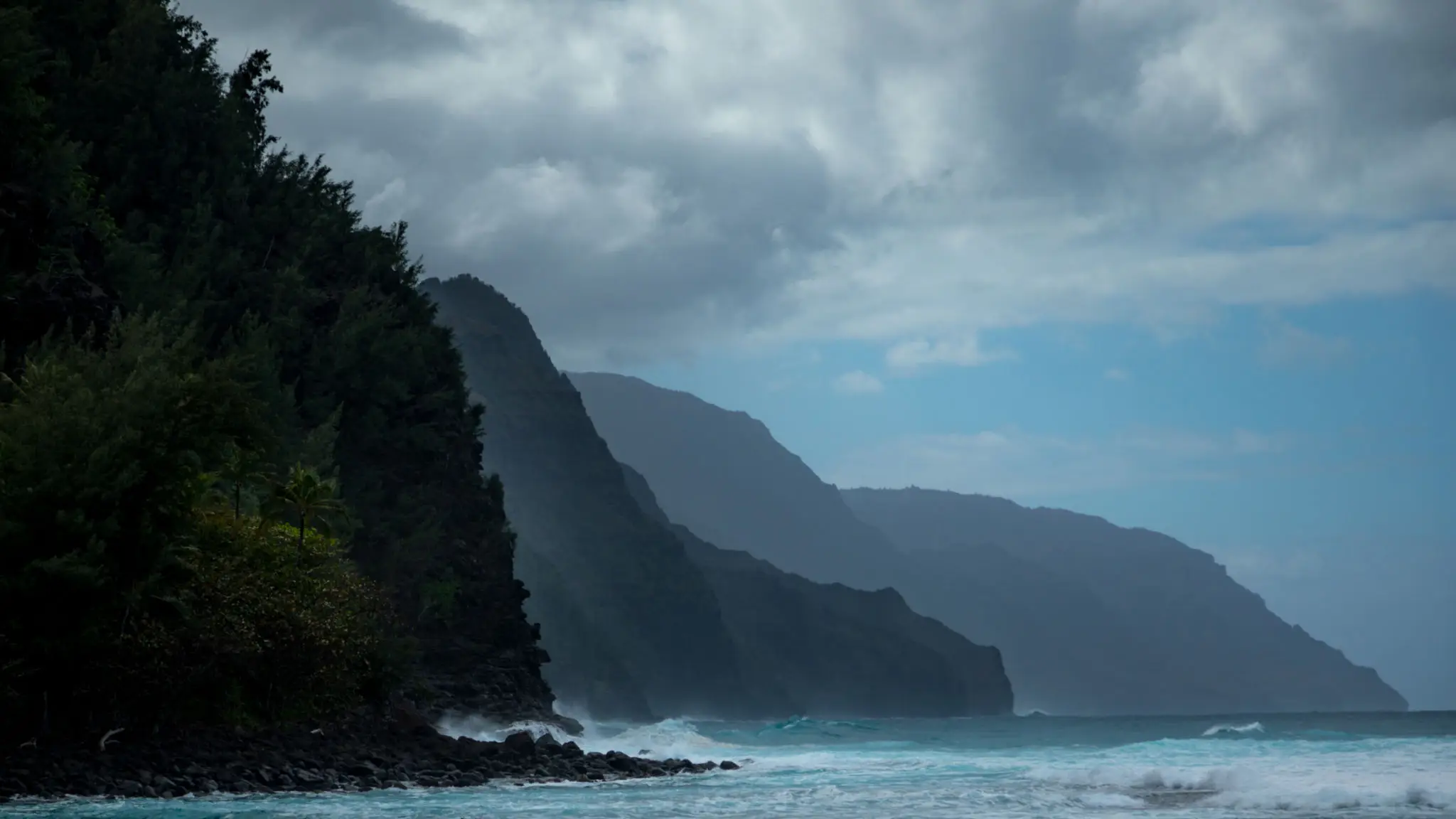 Misty cliffs rise above a turbulent ocean under a cloudy sky.