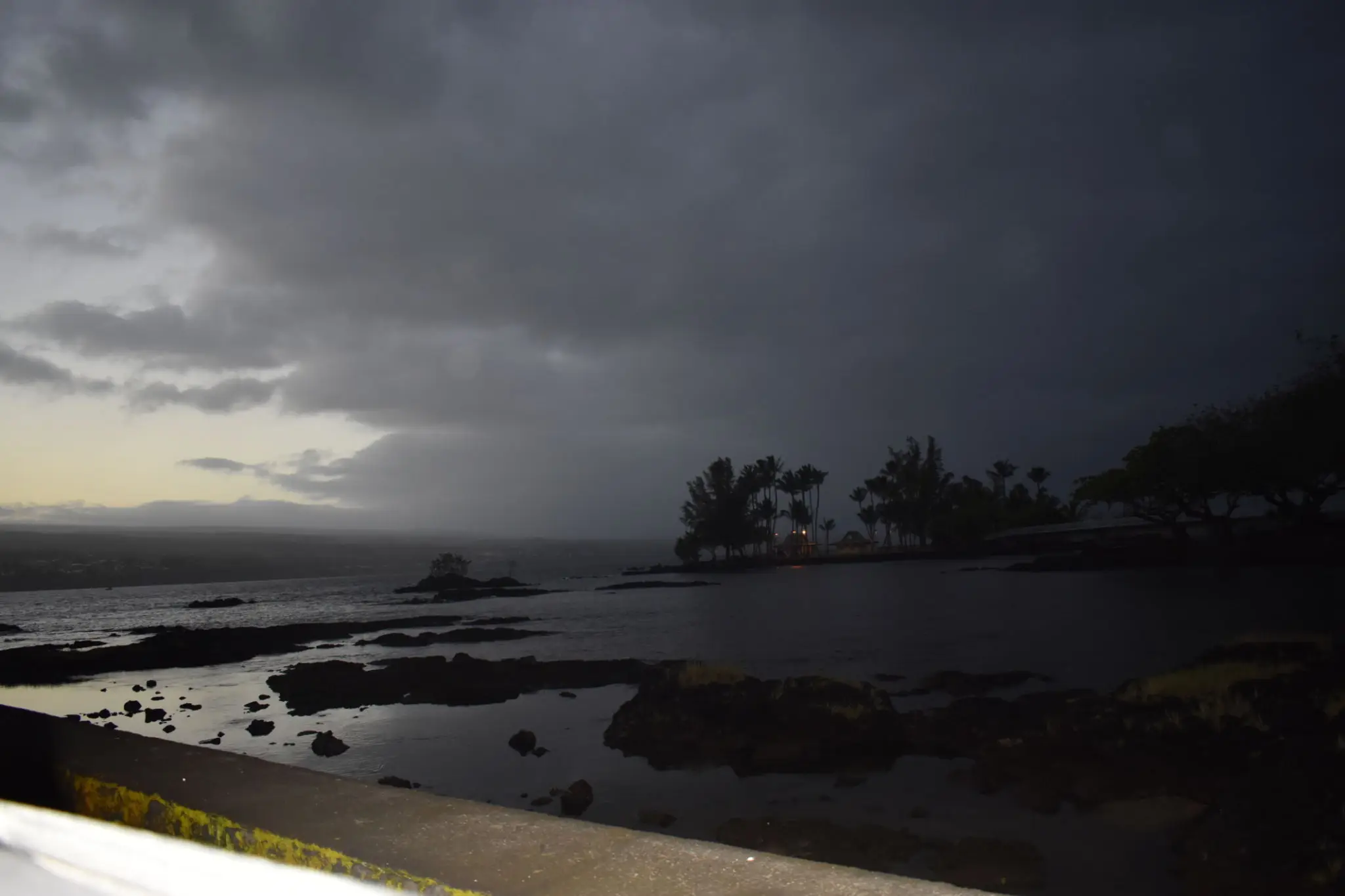 Dark storm clouds gather over a rocky shoreline with silhouetted trees.